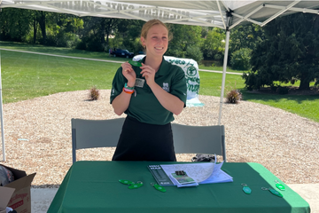 A person with blonde hair tied back, wearing a green polo shirt, smiling while standing under a canopy at an outdoor event. A table in front of them has informational materials and green keychains.