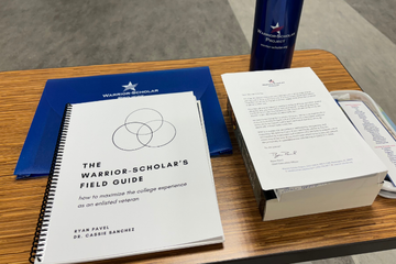 A desk with a booklet titled 'The Warrior-Scholar's Field Guide,' an informational pamphlet, a blue folder, and a blue water bottle, all items branded with the Warrior-Scholar Project logo.