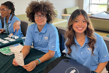 Three people sitting at a table, wearing matching blue polo shirts with a 'Mosaic 2024' logo. They are smiling and engaging with the camera; one person is holding a sheet of stickers, and another is pointing. The background includes a lounge area with seating and windows.