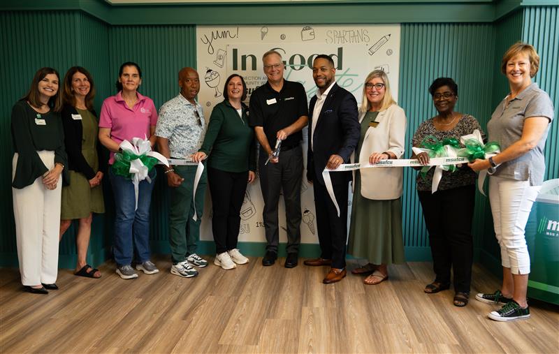 A group of people standing in a line, smiling, and holding a ribbon for a ribbon-cutting ceremony. They are in front of a wall with the text 'In Good Taste' and various drawings related to food. Several people hold green and white ribbons, and one person in the center holds a pair of scissors.