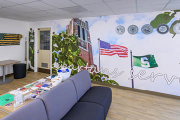 Interior of the Student Veterans Resource Center at Michigan State University, featuring a mural on the wall with the Spartan S flag and American flag, along with an image of Beaumont Tower. The room has a comfortable seating area with a table displaying various informational materials.