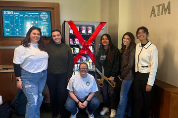 A group of six individuals stands beside a vending machine adorned with a large red ribbon and bow, symbolizing a ceremonial opening. The scene takes place indoors, with an ATM and bulletin board in the background. The group smiles, one member holding oversized ceremonial scissors.