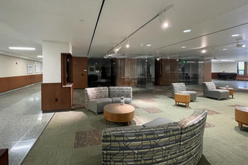 An open seating area inside a building with modern, cushioned chairs and circular tables. The room is softly lit with overhead lighting, and the floor is carpeted in a pattern of green and brown tones. Walls feature a mix of wood paneling and glass, creating a clean and inviting space for relaxation or study.