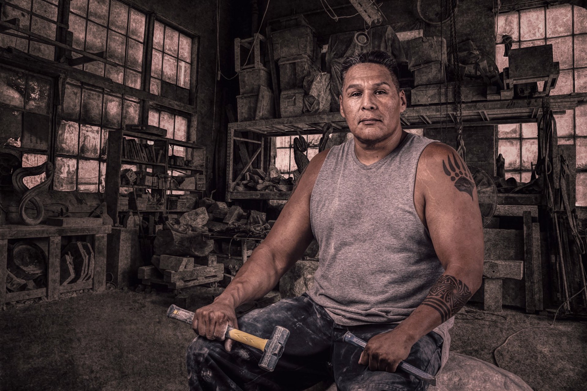 Jason Quingo sits in his sculpture workspace holding a hammer and chisel.