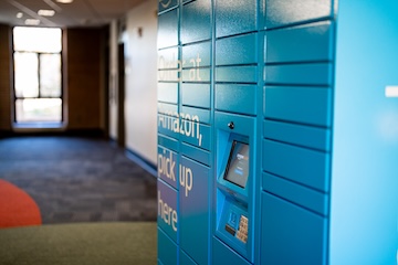 A bright blue Amazon locker system positioned in a quiet hallway with soft natural light coming through a window in the background. The locker is marked with text inviting users to pick up their packages.