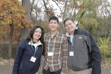 A family of three stand outside in front of trees on MSU's campus during Family Weekend, 2024.