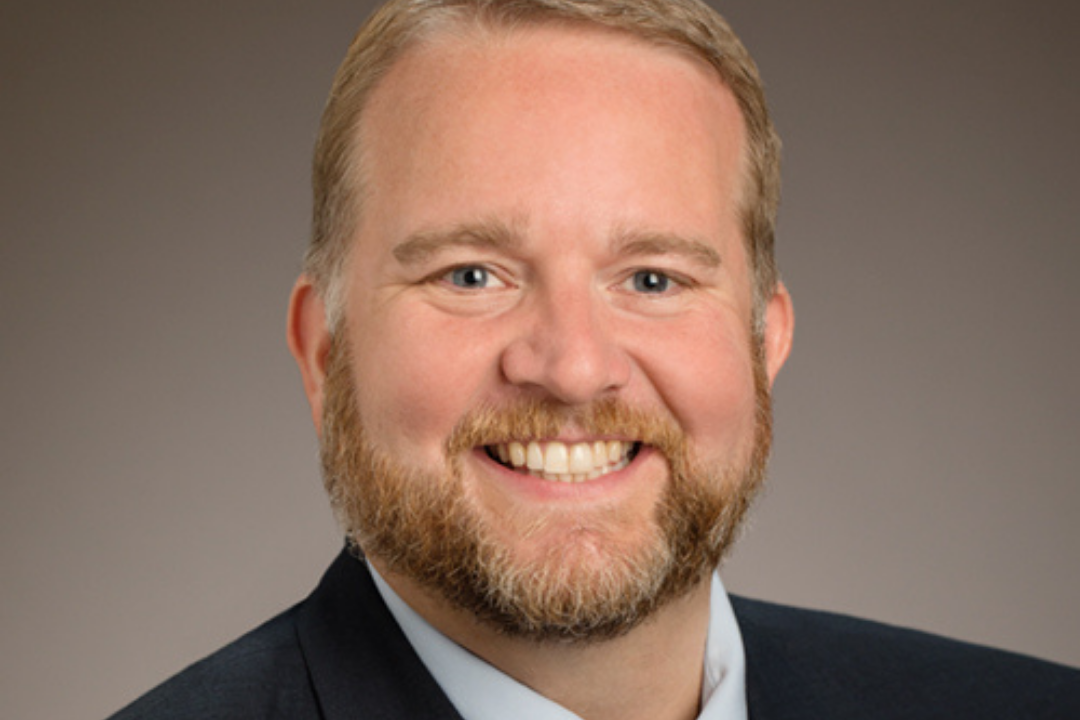 Smiling man in a suit and tie, with short blond hair and a full beard, posing in front of a neutral background.