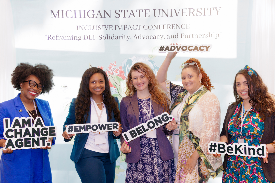 Five smiling individuals pose in front of a Michigan State University Inclusive Impact Conference backdrop. Each holds a motivational sign reading "I AM A CHANGE AGENT," "#EMPOWER," "I BELONG," "#ADVOCACY," and "#BeKind."
