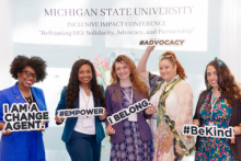 A group of five individuals smiling and holding signs with positive messages during the Michigan State University Inclusive Impact Conference. The signs read: 'I AM A CHANGE AGENT,' '#EMPOWER,' 'I BELONG,' and '#BeKind.' The backdrop displays the event's theme: 'Reframing DEI: Solidarity, Advocacy, and Partnership,' with the hashtag #ADVOCACY prominently featured.