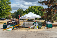 A plastic and metal recycling collection point set up under a white tent at Michigan State University, with bins labeled for different types of recyclables. The area is marked with a 'Campus Entrance' sign and surrounded by trees and a road in the background.