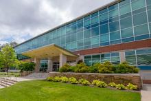 The Student Services Building on campus, featuring a modern facade with large glass windows, a covered entrance, and landscaped greenery in the foreground under a partly cloudy sky.