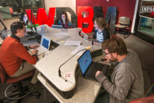 Students collaborating at the WDBM Impact 89FM radio station at Michigan State University. The group, gathered around a broadcast desk with laptops and notes, showcases hands-on experience in media production and teamwork.