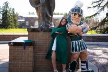 SFC student employee, Lindsey, stands with her back against Sparty's, both of them with their arms confidently crossed, below the Spartan statue.