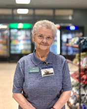 Longtime Culinary Services team member Clara Kent stands in front of a food display wearing a blue shirt and glasses. 