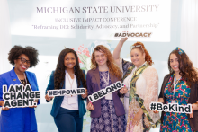 Five smiling individuals pose in front of a Michigan State University Inclusive Impact Conference backdrop. Each holds a motivational sign reading "I AM A CHANGE AGENT," "#EMPOWER," "I BELONG," "#ADVOCACY," and "#BeKind."