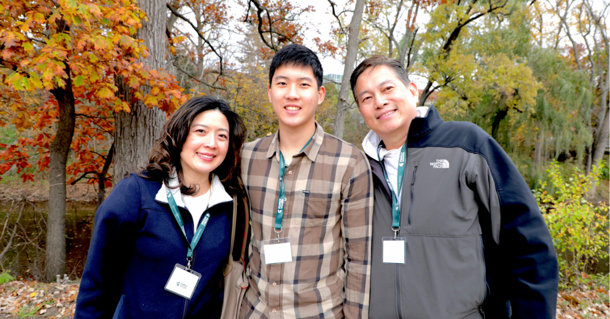 Three people standing outdoors in a wooded area with fall foliage. They are smiling and wearing lanyards with ID badges. One person is wearing a plaid shirt, and the other two are wearing jackets.